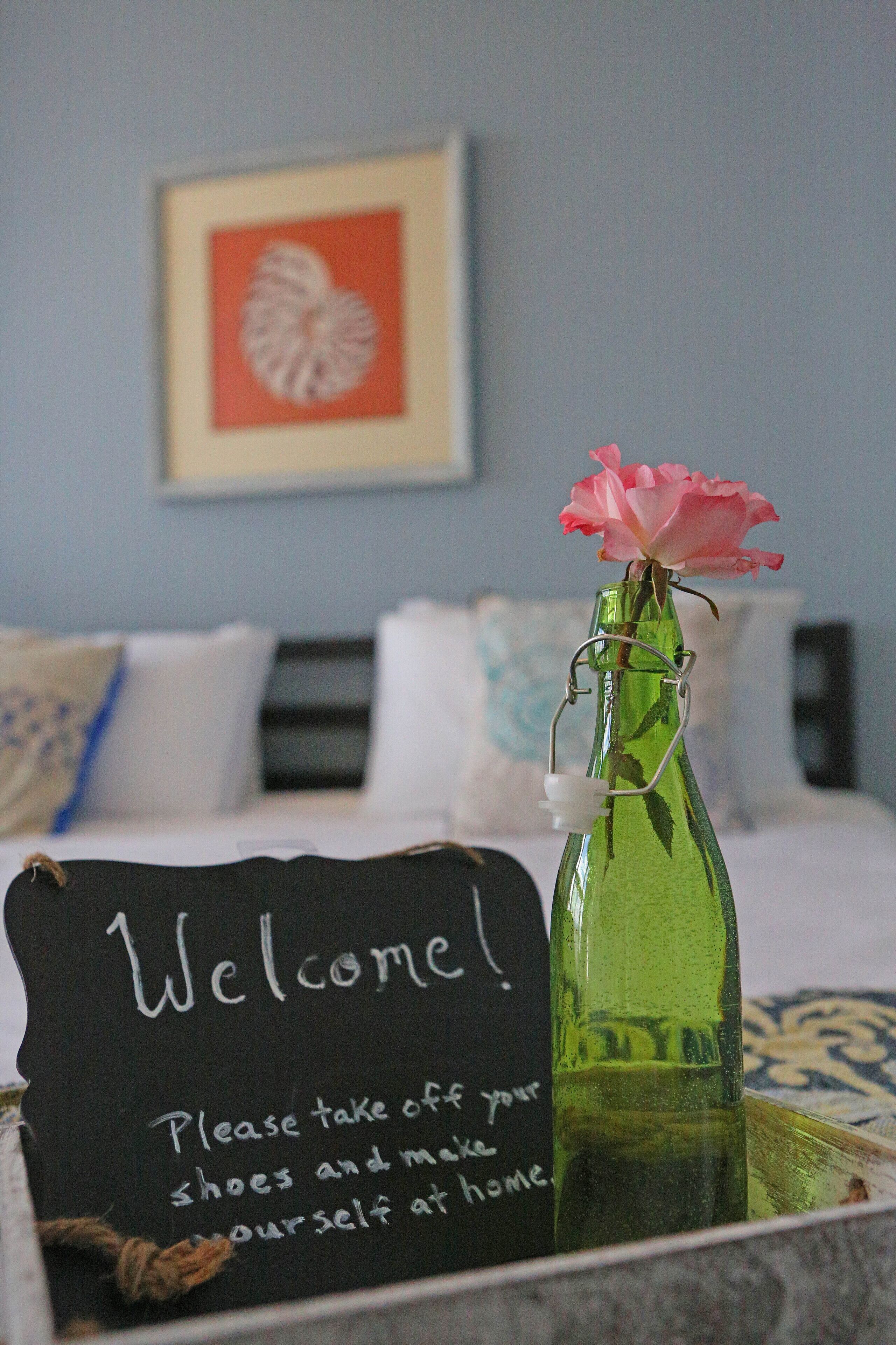 A bed with a pillow, a bedside table with a green bottle and a sign that says "Welcome! Please take off your shoes and make yourself at home."