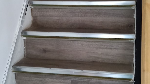 Un escalier en bois avec des rampes en métal, menant à une pièce dont le mur est blanc et qui contient une table noire.