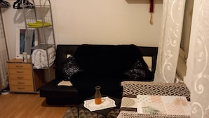 Une pièce avec un canapé noir, une petite table sur laquelle se trouve une bouteille, des coussins à motifs, une étagère en métal avec des vêtements et un rideau à motifs.