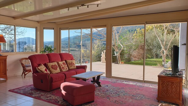 Ein geräumiges Wohnzimmer mit einem roten Sofa, einem Couchtisch und einer großen Schiebeglastür, die auf einen Balkon mit Blick auf Bäume und Berge führt.
