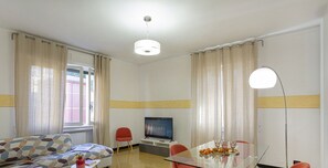 A modern living room with a sofa, a glass dining table with red chairs, a TV, and a lamp.
