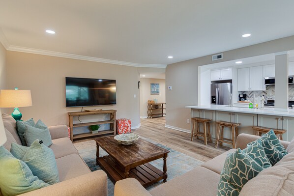 A modern living room with a sofa, coffee table, and TV. A kitchen with white cabinets and a stainless steel refrigerator is visible in the background. - Newly Renovated! Water View 3 Bed/2 BA Villa @ South Beach & Salty Dog, Sleeps 8 (Hilton Head Island)