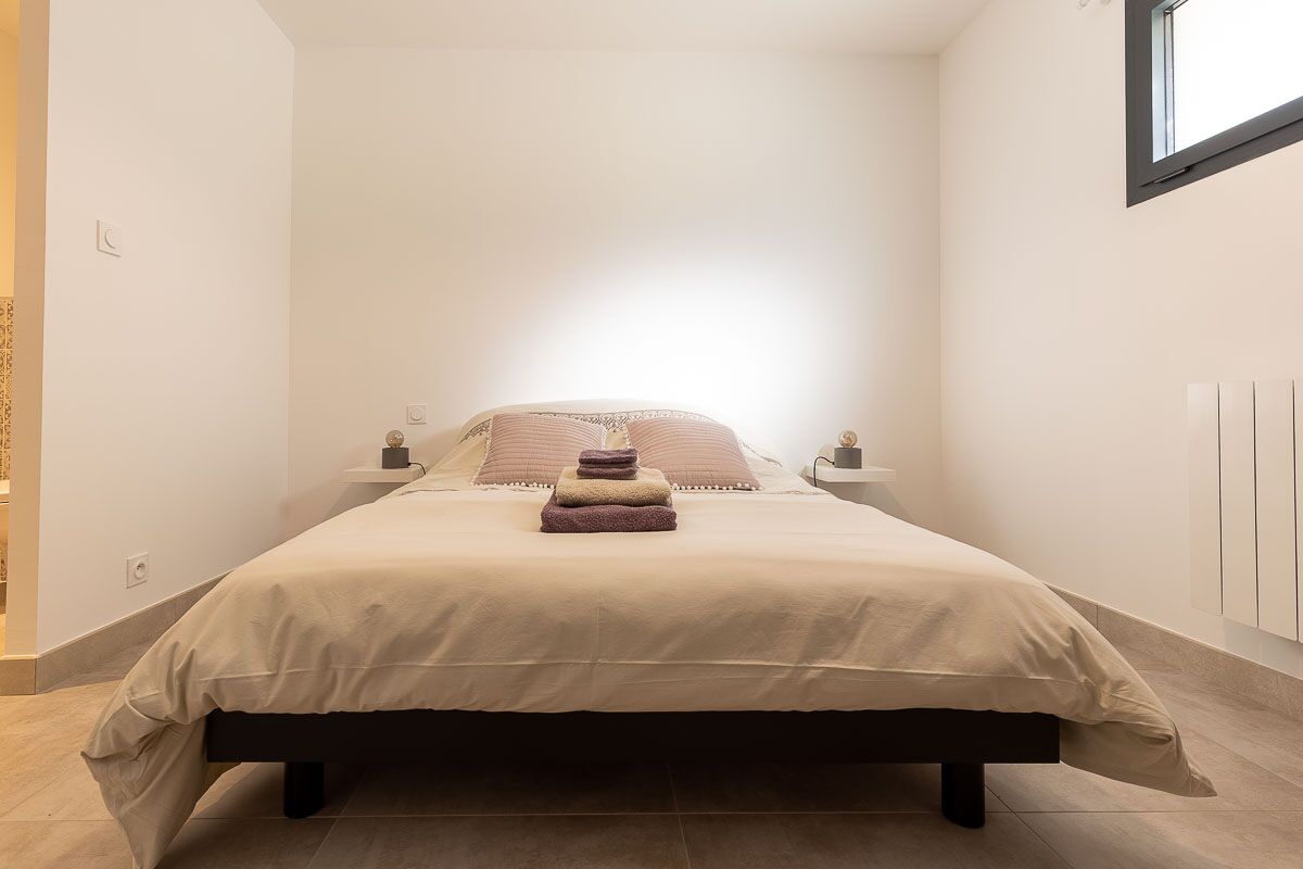 Una cama bien hecha con una colcha color crema y almohadas, una mesita de noche con una lámpara, y una ventana que deja entrar luz natural.