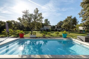 A rectangular swimming pool with a clear blue water surface, surrounded by a concrete deck. There are potted plants and a grassy area with trees in the background.