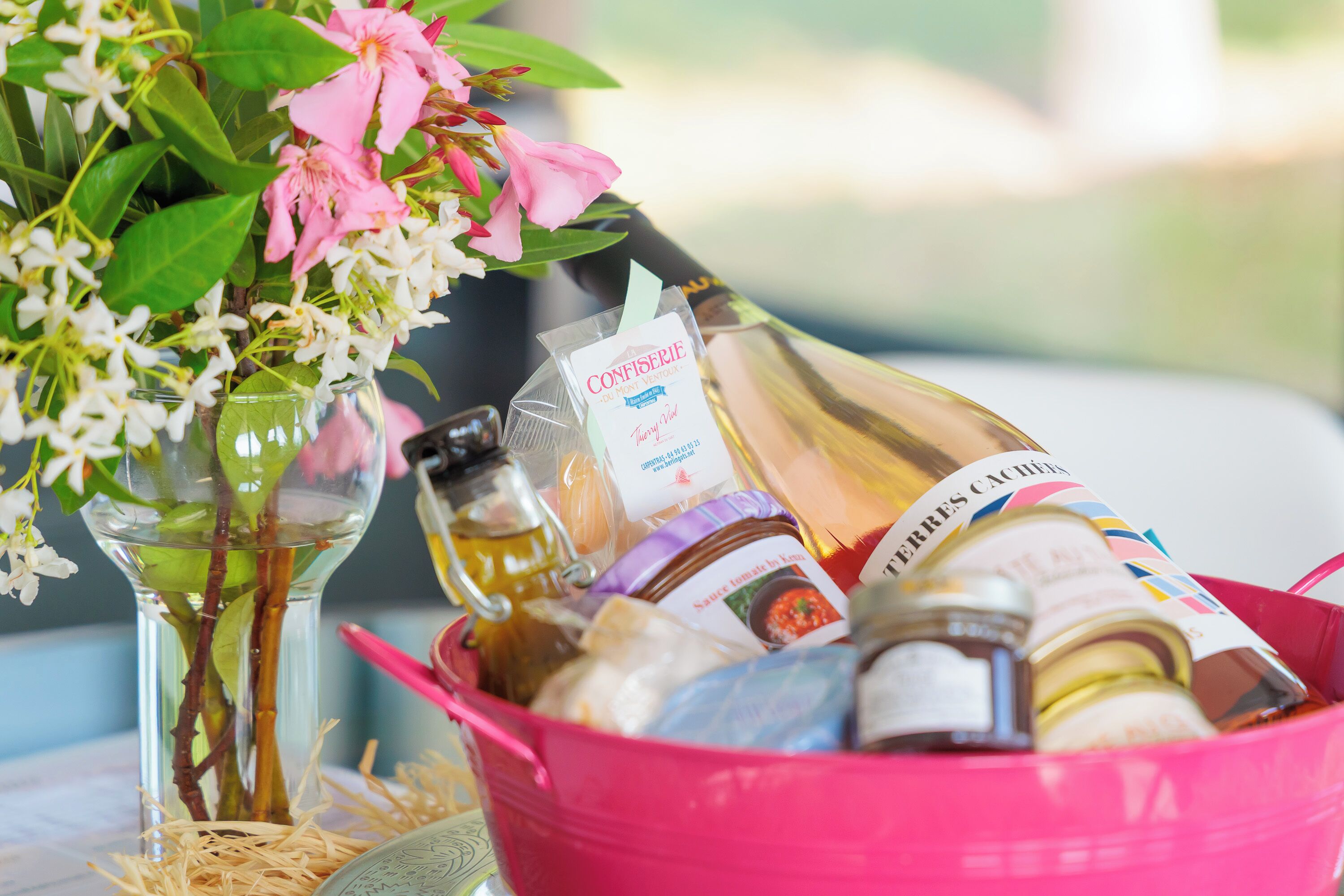 Un seau rose rempli de différentes bouteilles, dont une bouteille de vin et un pot de confiture, est posé sur une table, à côté d’un vase de fleurs.