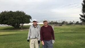 Two people standing on a golf course with a tree and a fence in the background.