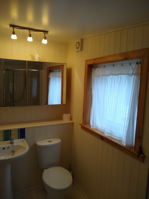 Un baño con lavamanos, inodoro y espejo. Hay una ventana con cortina y una zona de ducha con puerta de vidrio.