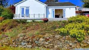 Une maison blanche à deux étages, avec un mur de pierre à l’avant, une terrasse en bois et un garage dans lequel une voiture est garée.