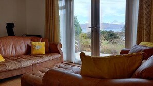 Un salon avec un canapé en cuir marron, deux coussins jaunes et une grande fenêtre donnant sur des montagnes et un lac.