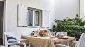 A dining area with a table set for two, white chairs, a window with shutters, and a potted plant. - Apartment Brezza by Interhome (Scicli)