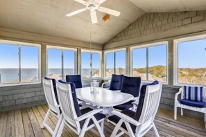 Un espace repas abrité, avec une table et des chaises, de grandes fenêtres donnant sur la mer et un ventilateur de plafond.