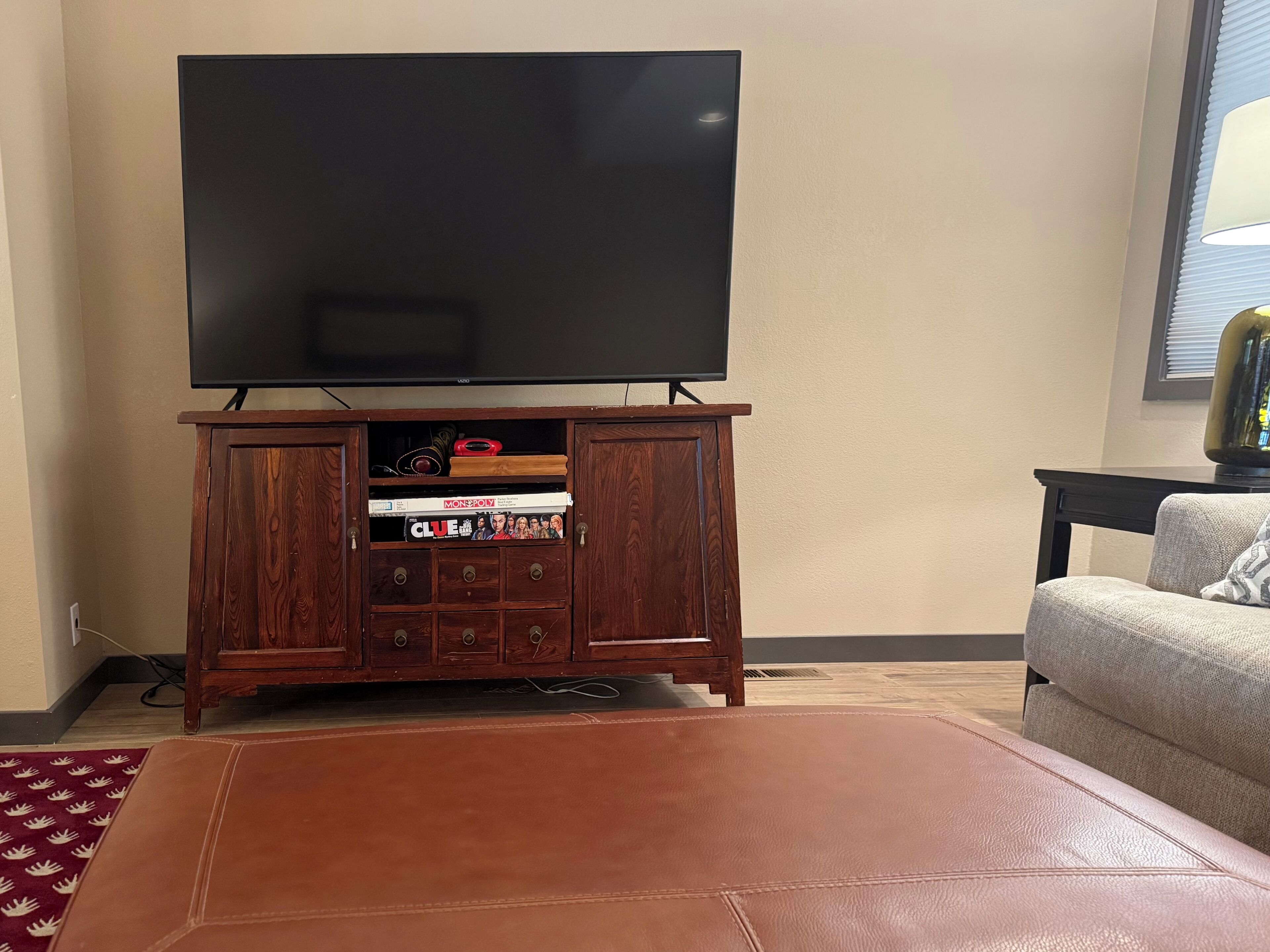 Una sala de estar con televisor de pantalla plana montado en un mueble de madera, un reposapiés de cuero y un sofá.