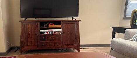 Una sala de estar con televisor de pantalla plana montado en un mueble de madera, un reposapiés de cuero y un sofá.