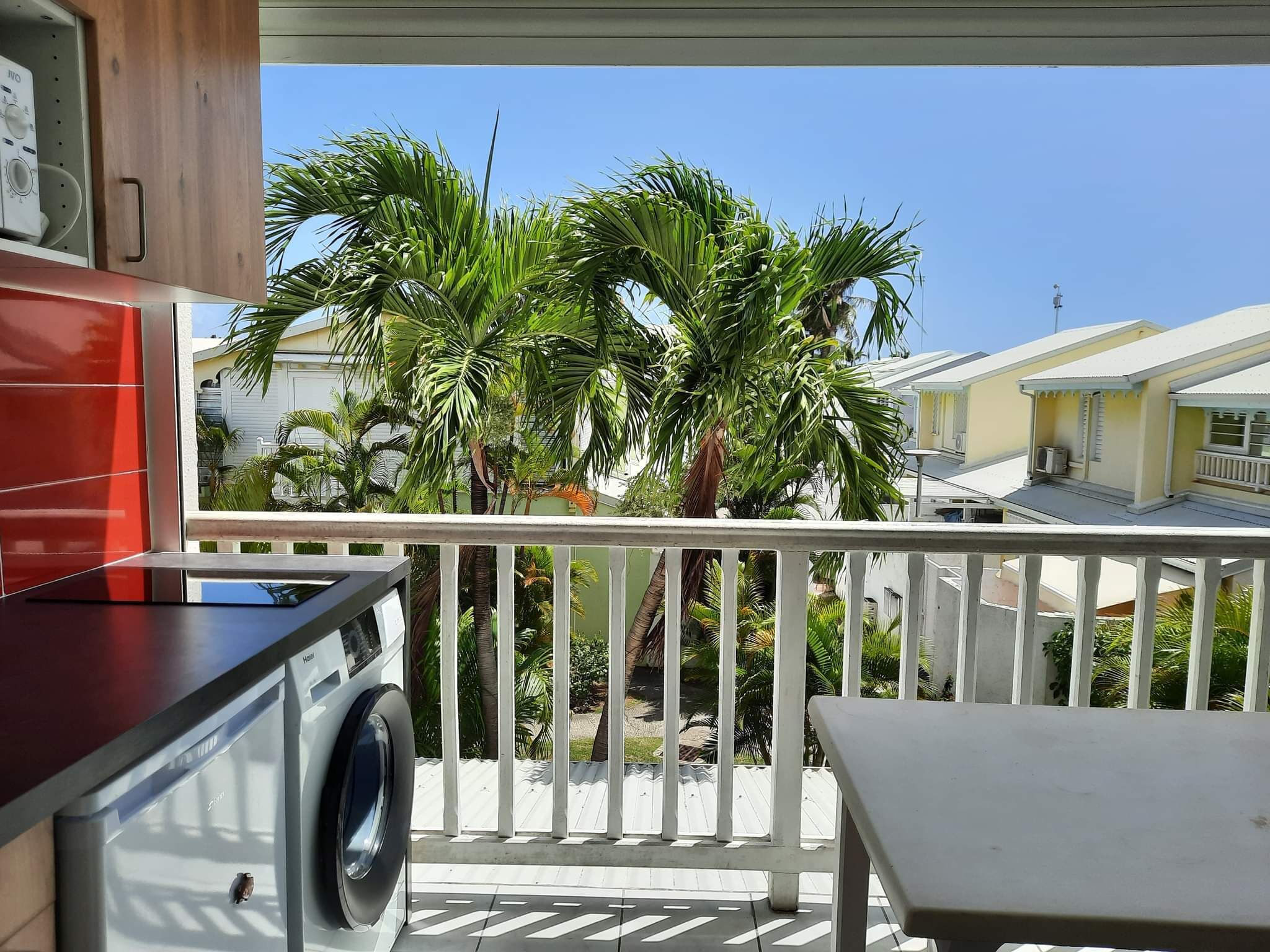 A balcony with a washing machine, a table, and a view of palm trees and neighboring houses.