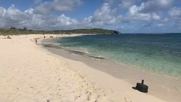 Ein sandiger Strand mit klarem türkisblauem Wasser, einigen Personen und einer in der Ferne liegenden Felsformation.