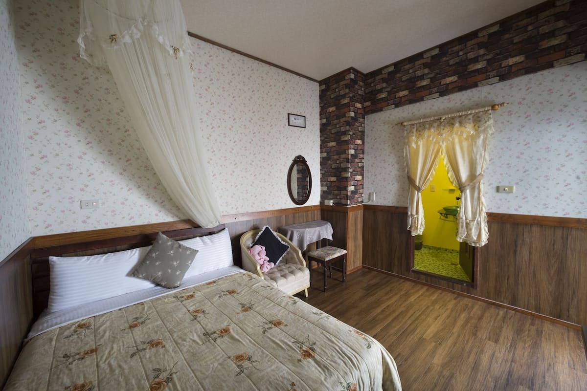 A bedroom with a bed, a chair, a small table, a mirror, and a window with curtains.