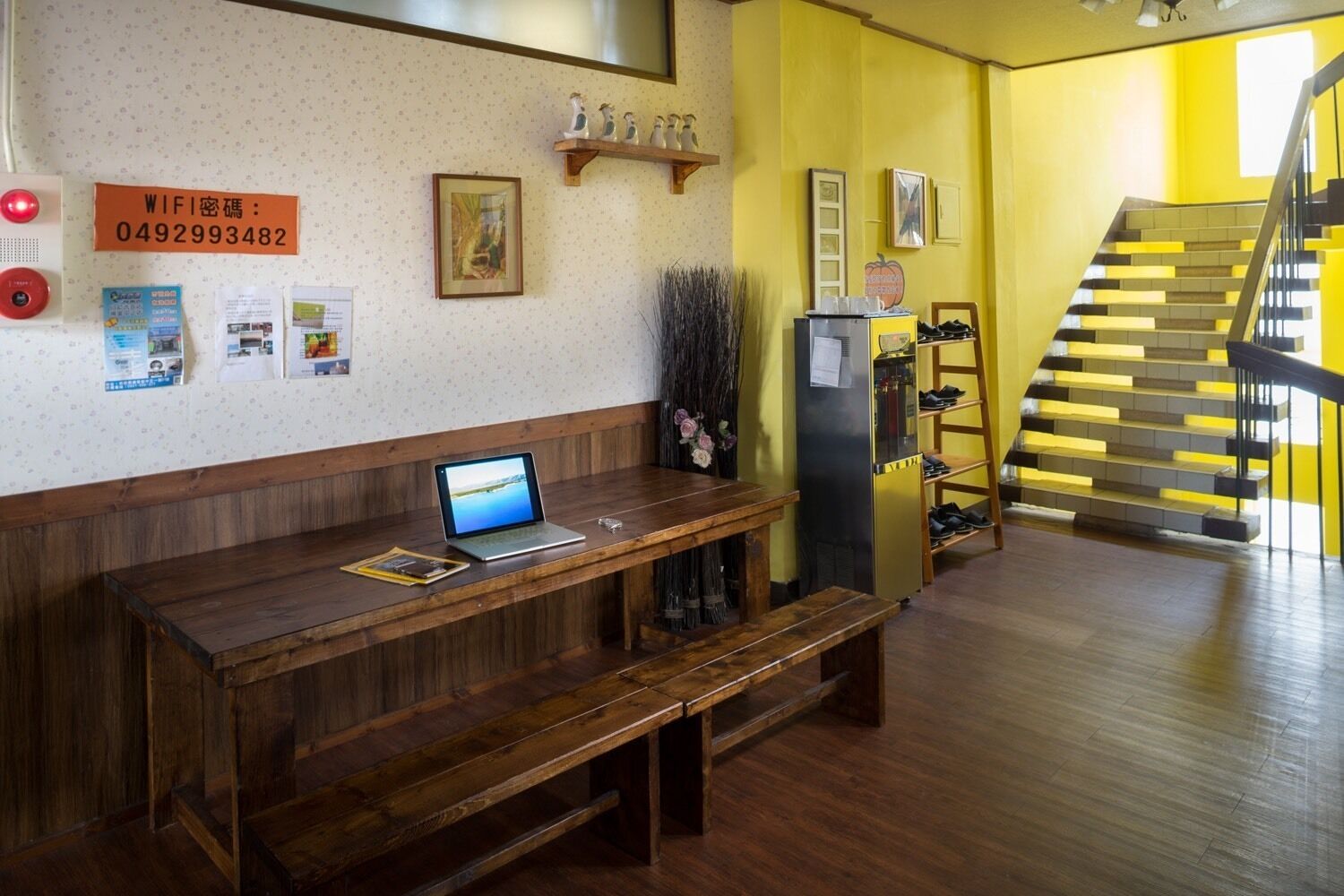A reception area with a wooden bench, a laptop, a refrigerator, and a staircase with yellow walls.