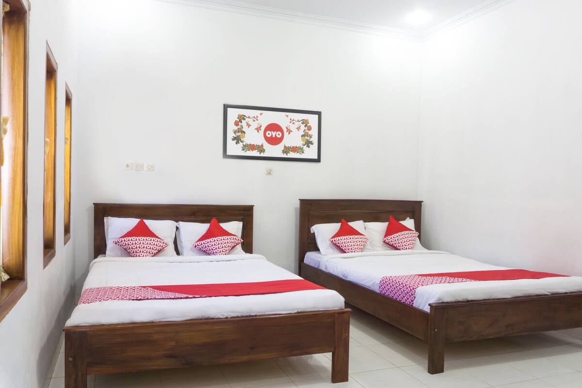 Two wooden beds with white bedding and red accents, a framed picture on the wall, and a window with wooden frames.