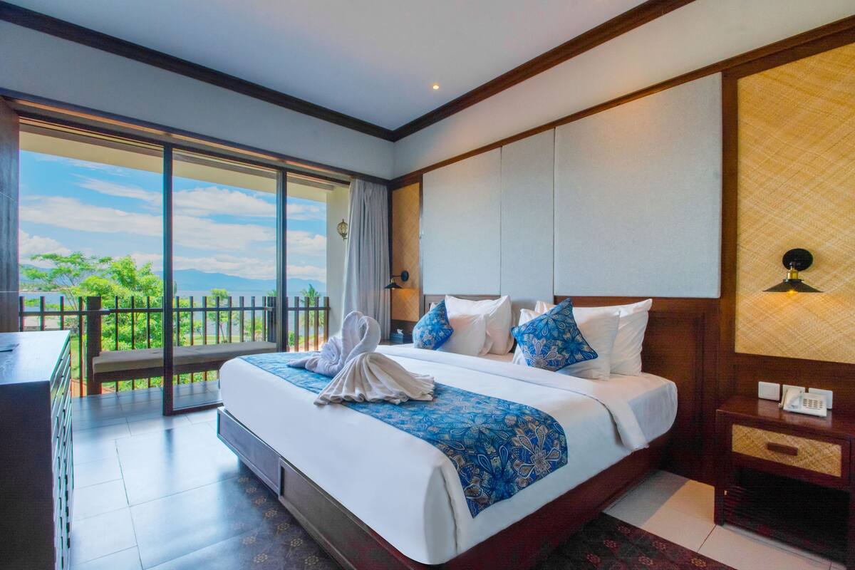 A hotel room with a large bed, a desk, and a balcony with a view of greenery and mountains.