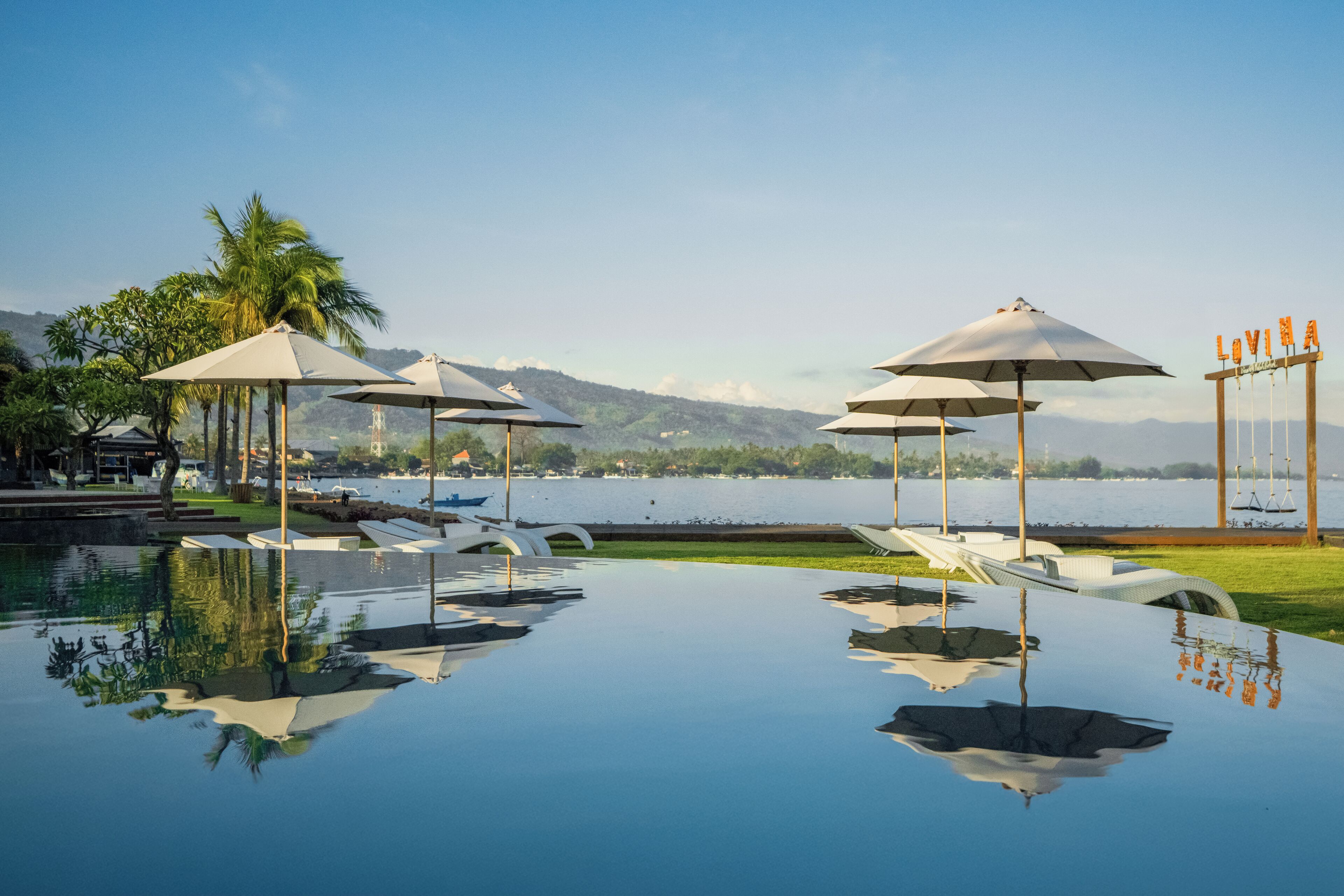 An infinity pool with lounge chairs and umbrellas, a view of the lake, and a sign with the name 'LOVINA'.