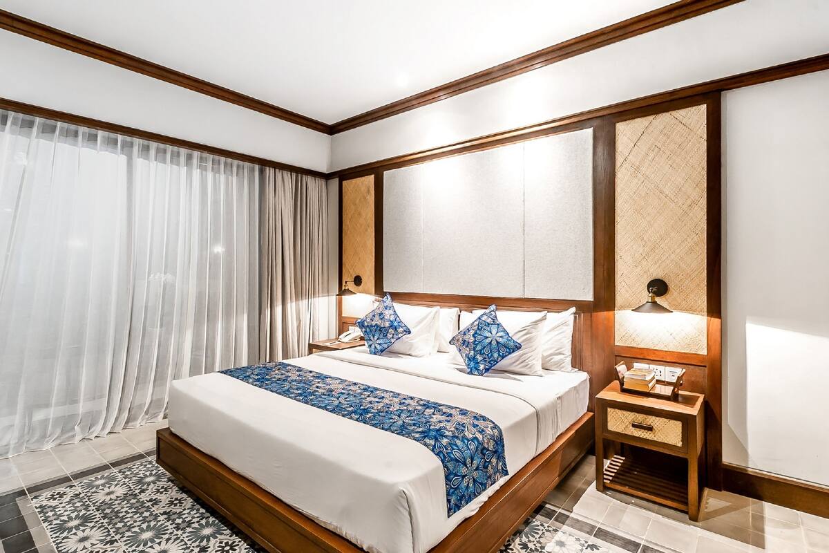 A hotel room with a large bed, wooden headboard, a bedside table with a lamp, and a patterned rug.