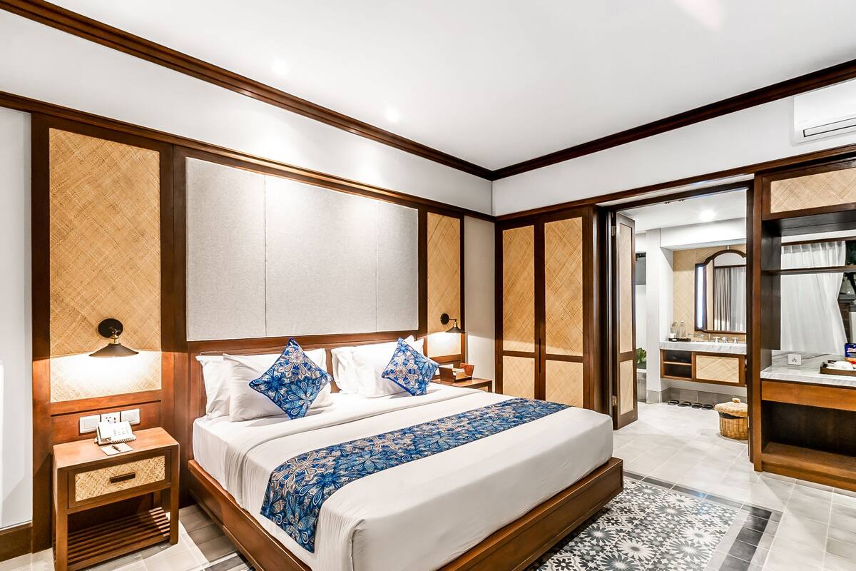 A modern hotel room with a large bed, wooden headboard, bedside tables with lamps, and a bathroom visible through an open door.