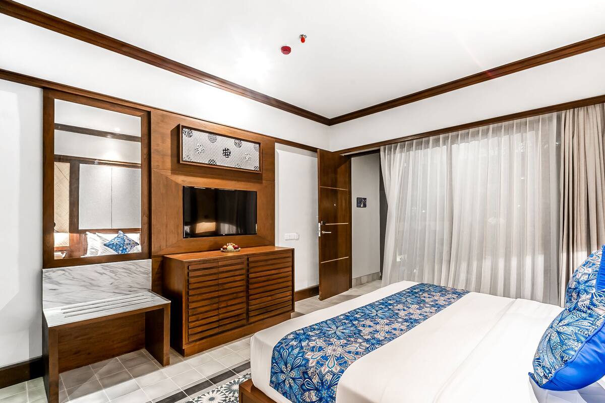 A modern hotel room with a large bed, a wooden dresser, a vanity with a mirror, and a sliding door closet.