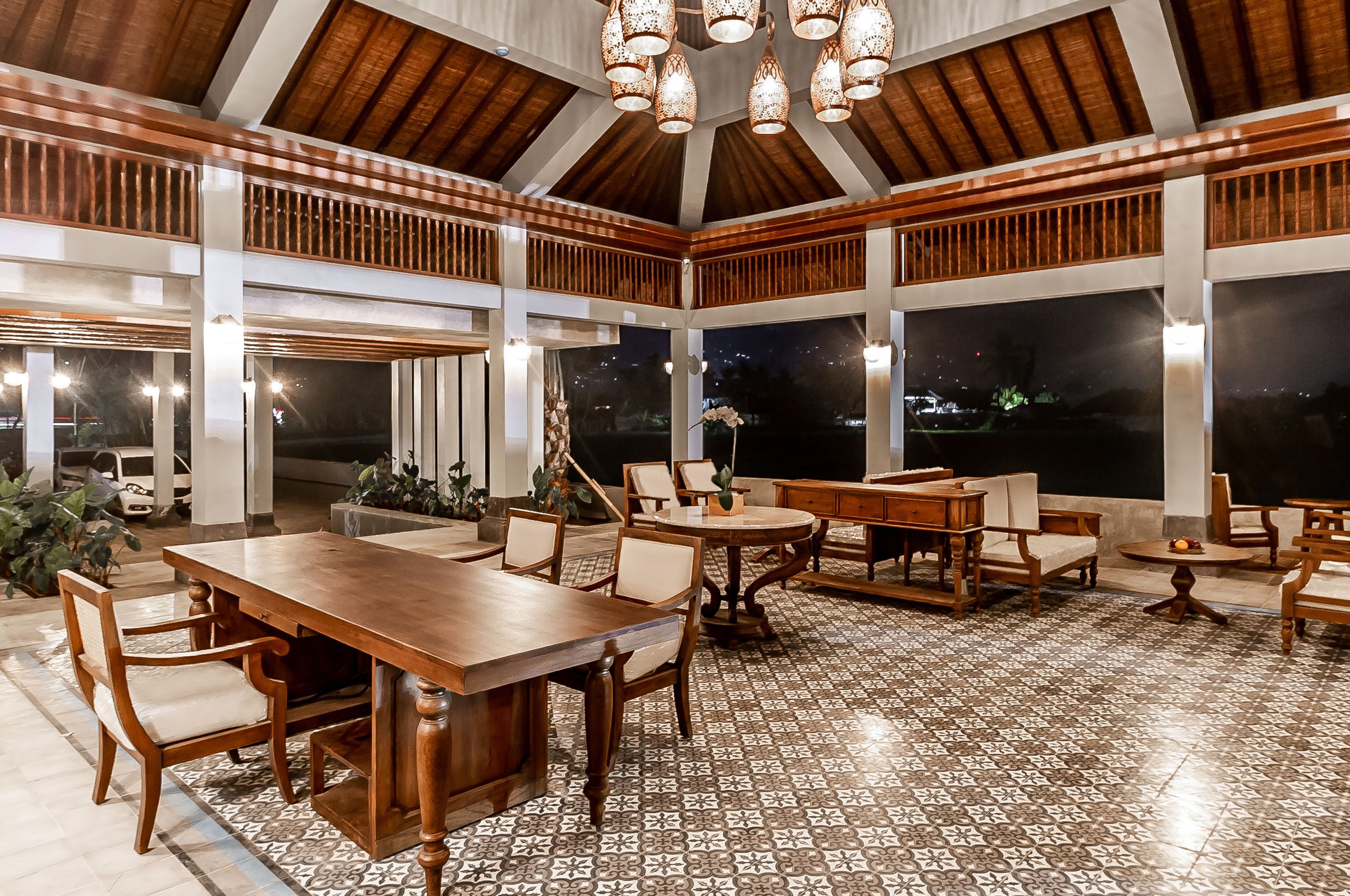 A spacious dining area with a high ceiling and large windows, featuring a wooden dining table and chairs, surrounded by potted plants.