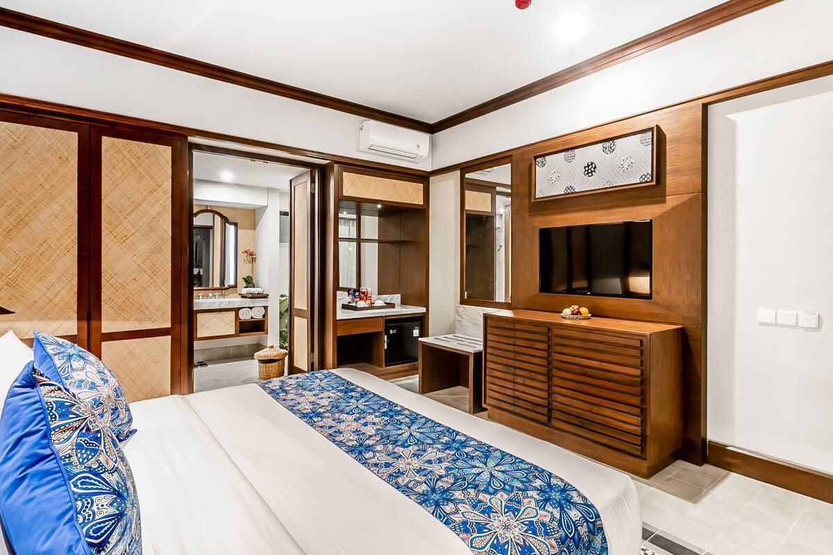 A modern hotel room with a large bed, a wooden dresser, a TV mounted on the wall, and a bathroom visible through an open door.