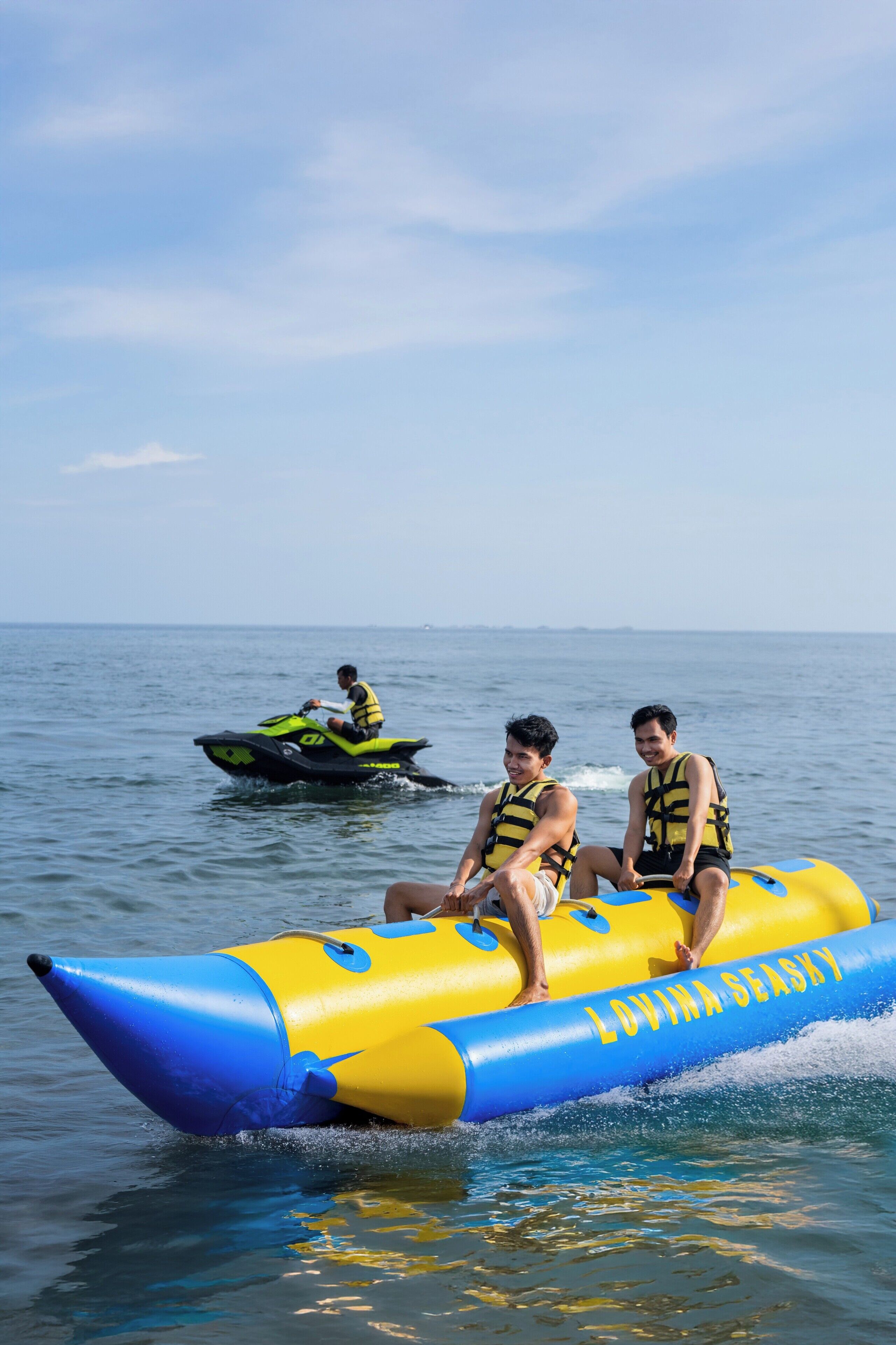 Two people on a yellow and blue inflatable raft with the text "LOVINA SEASHY" on it. Another person is on a green jet ski in the background.