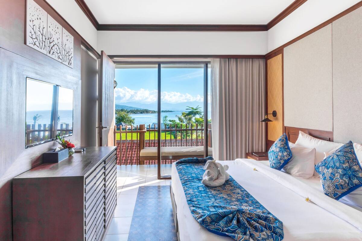 A hotel room with a large bed, a TV, a desk, and a balcony with a view of the sea.