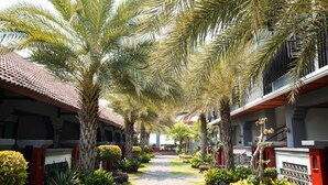 Un sentier bordé de palmiers et de différentes plantes, qui mène à un bâtiment doté d’un toit en tuiles et de détails en rouge.