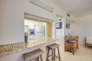 A kitchen with granite countertops, a bar stool, a wicker side table, and a view into the dining area.