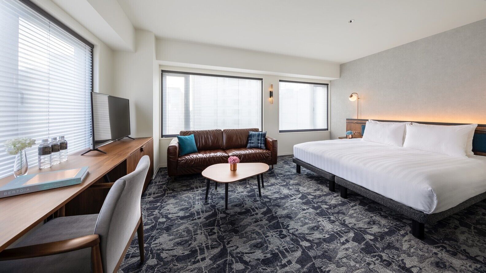 A hotel room with a bed, a desk, a sofa, a coffee table, and two windows with blinds.