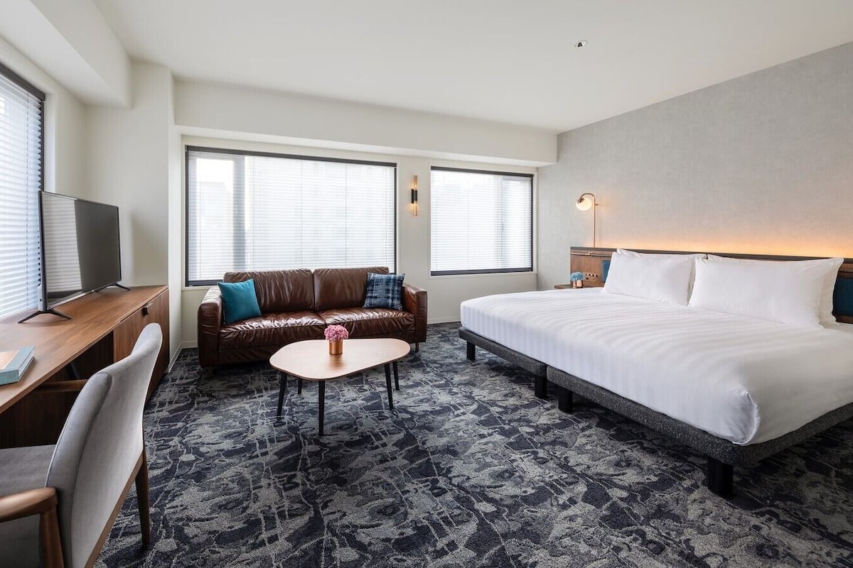 A hotel room with a bed, a desk, a sofa, a coffee table, and two windows with blinds.