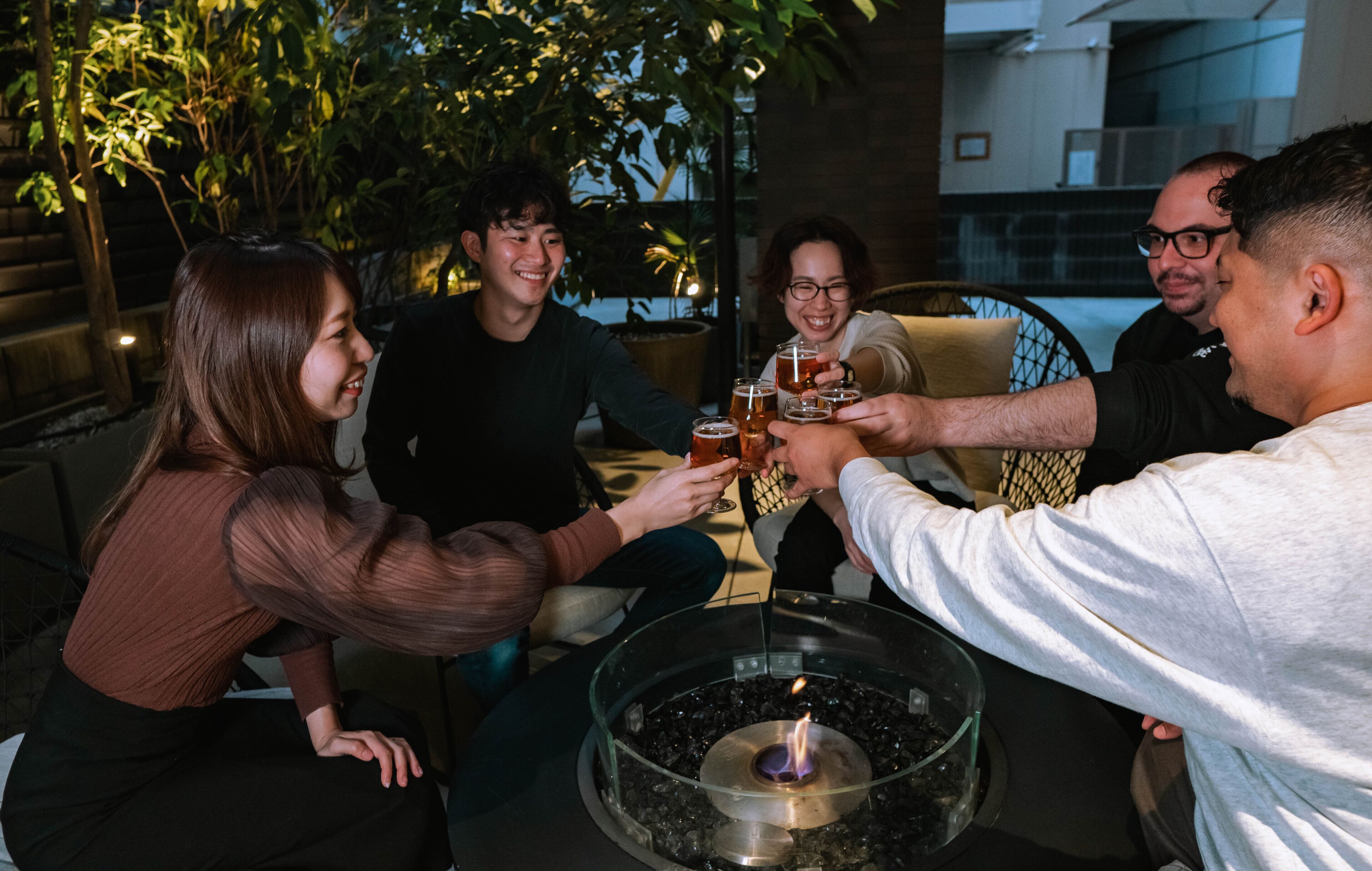 Un grupo de personas brindando con bebidas alrededor de una mesa de vidrio con una chimenea central.