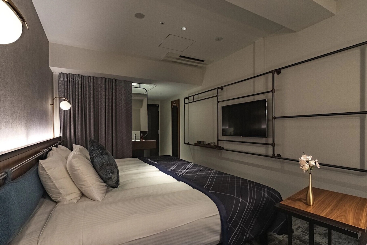 A hotel room with a bed, a TV, a small table with a vase, and a window with curtains.