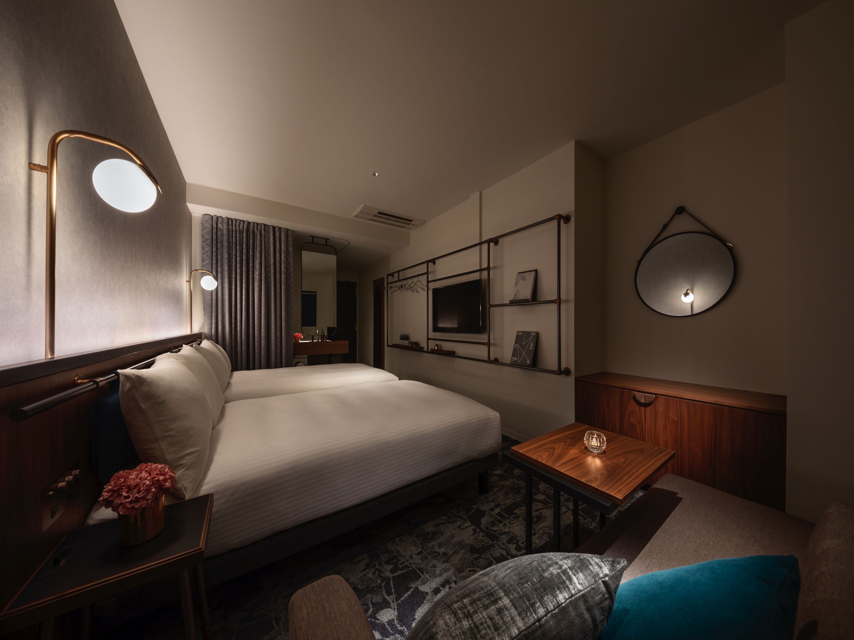 A hotel room with a large bed, bedside table, a small wooden table, a mirror, a TV mounted on the wall, and a wall-mounted shelf with items.