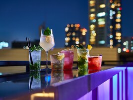 Une variété de cocktails colorés sont présentés sur le comptoir d’un bar, avec une vue sur les gratte-ciel de la ville en arrière-plan.