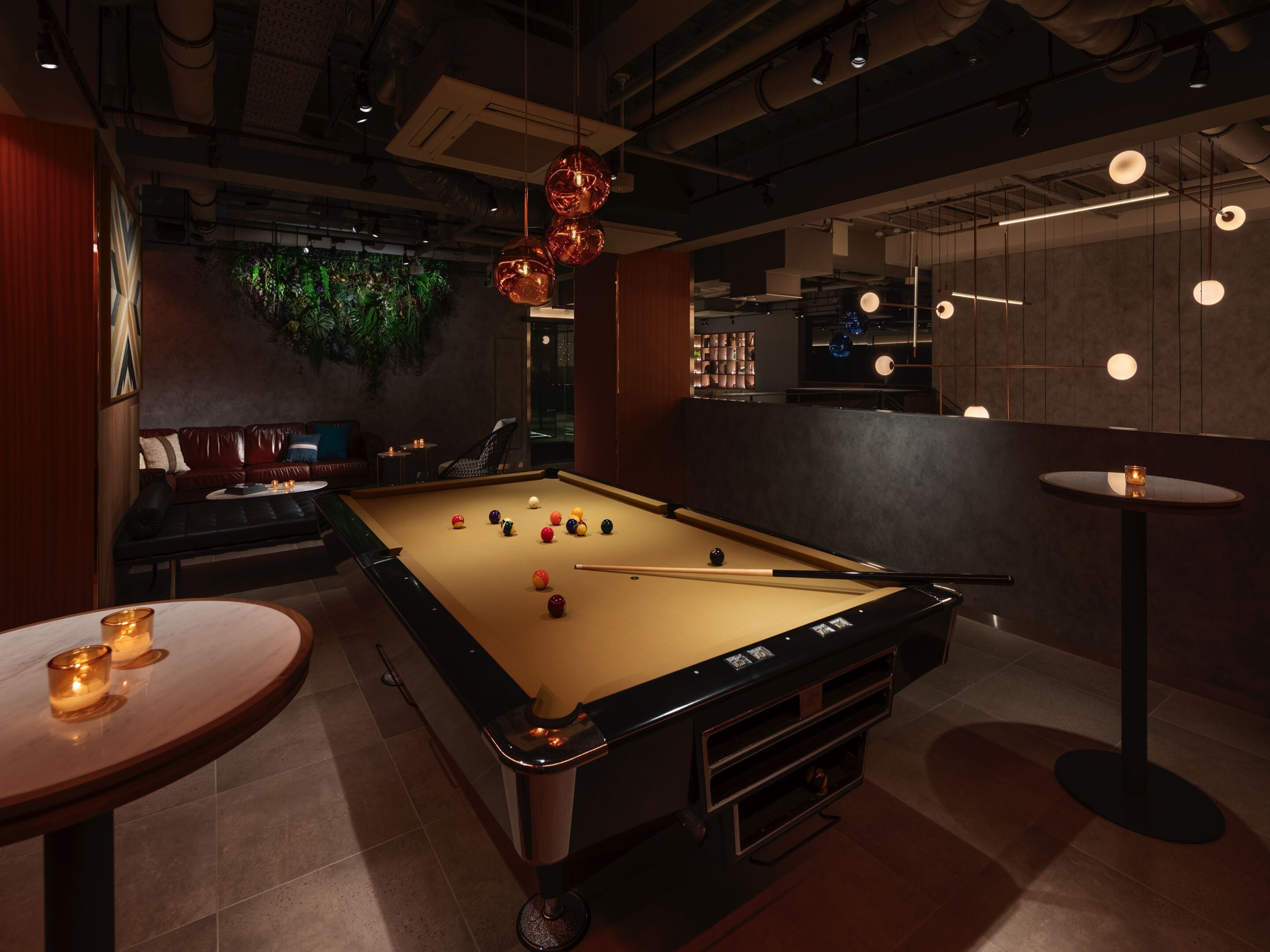 A billiards table in a dimly lit room with ambient lighting, a green wall with plants, and a bar area with shelves and lighting fixtures.