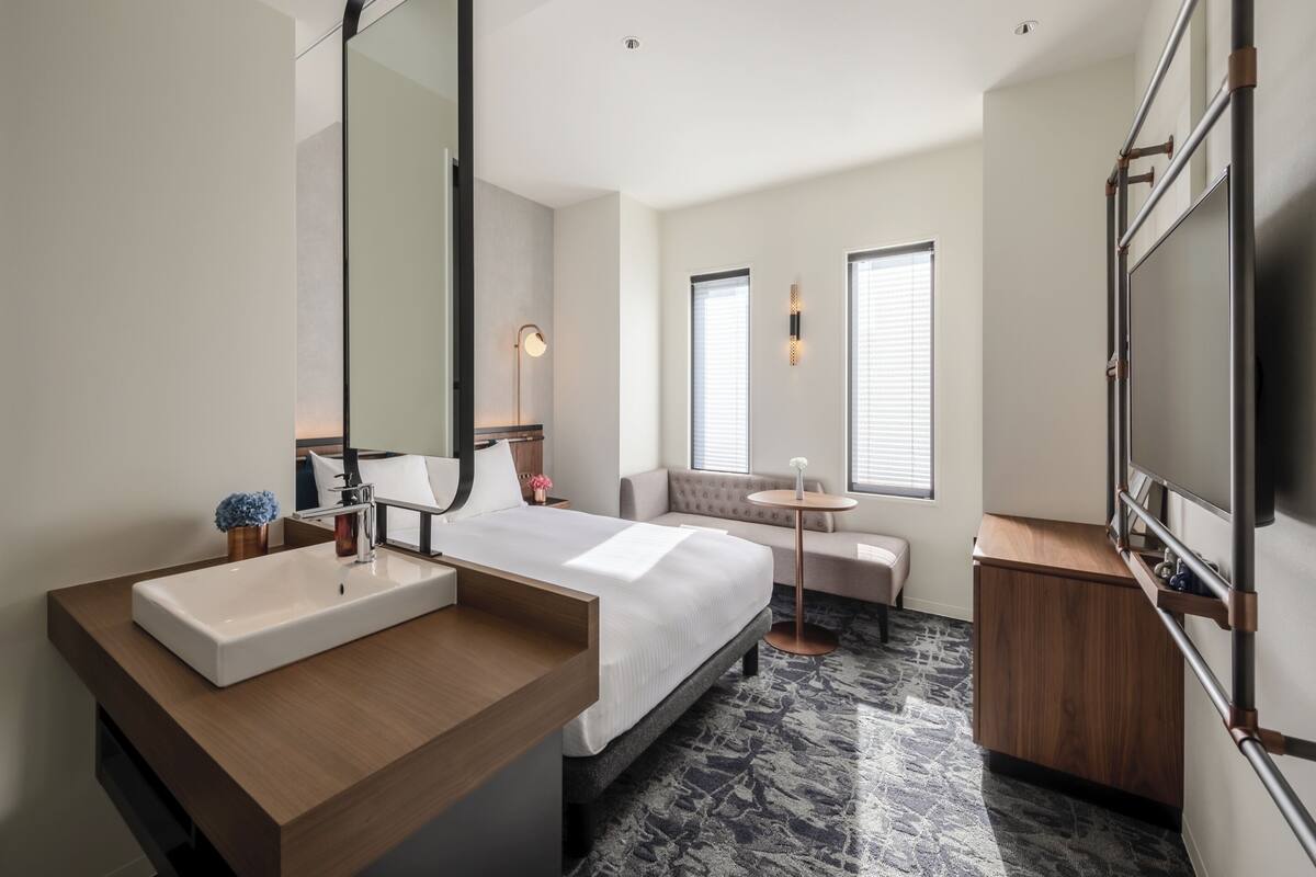 A modern hotel room with a large bed, a bedside table with a sink, a mirror, a small round table with a vase, and a sofa.