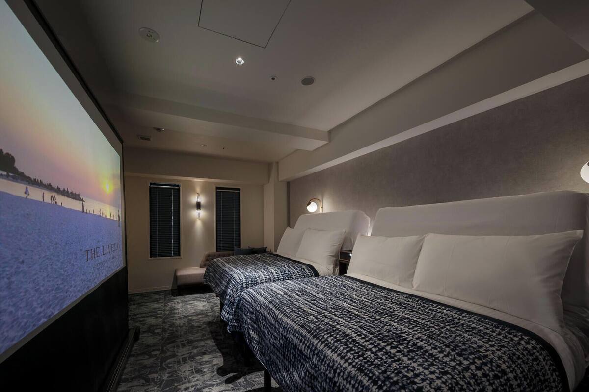 A hotel room with a large bed, a smaller bed, a TV screen, and a projector displaying a scenic image.