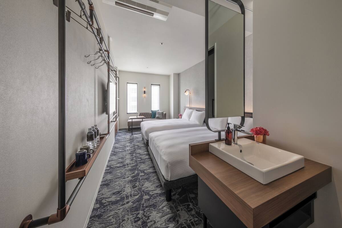 A hotel room with a large bed, a smaller bed, a bathroom with a sink and mirror, and a bench with a view of the hallway.