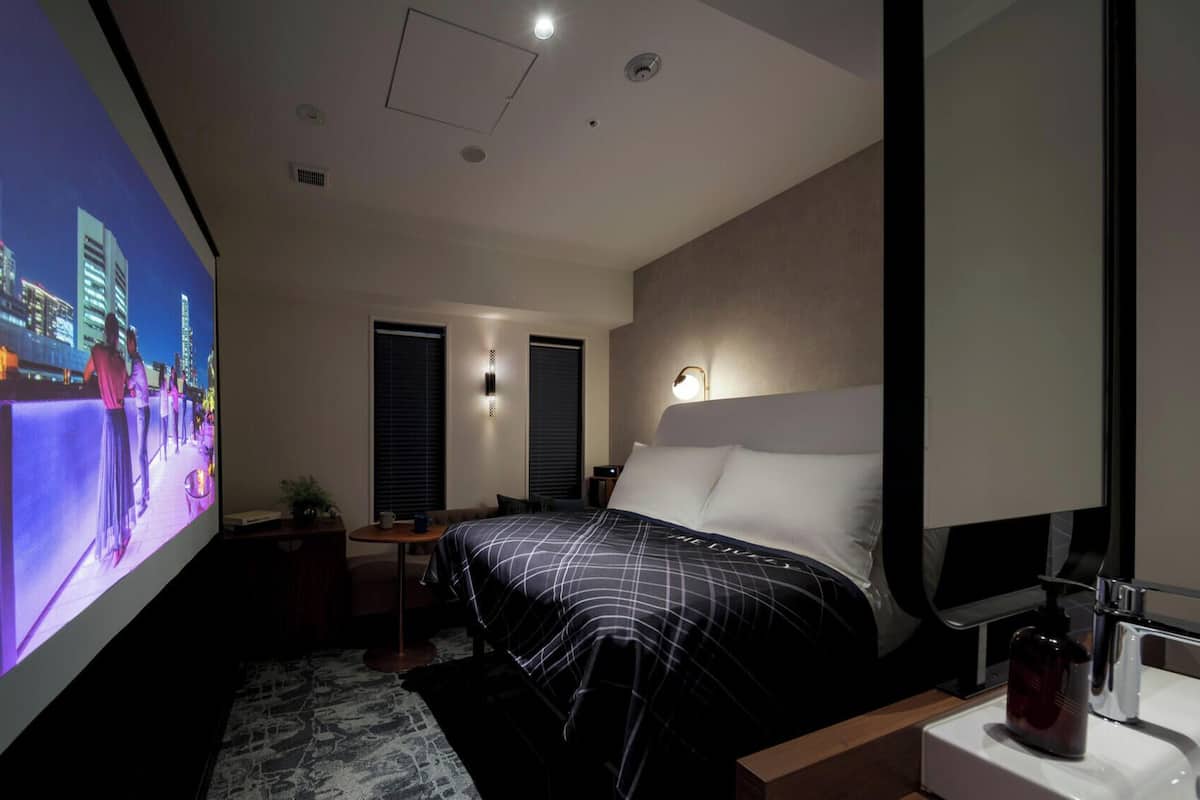 A modern hotel room with a large bed, a bedside table, a TV, and a window with blinds.