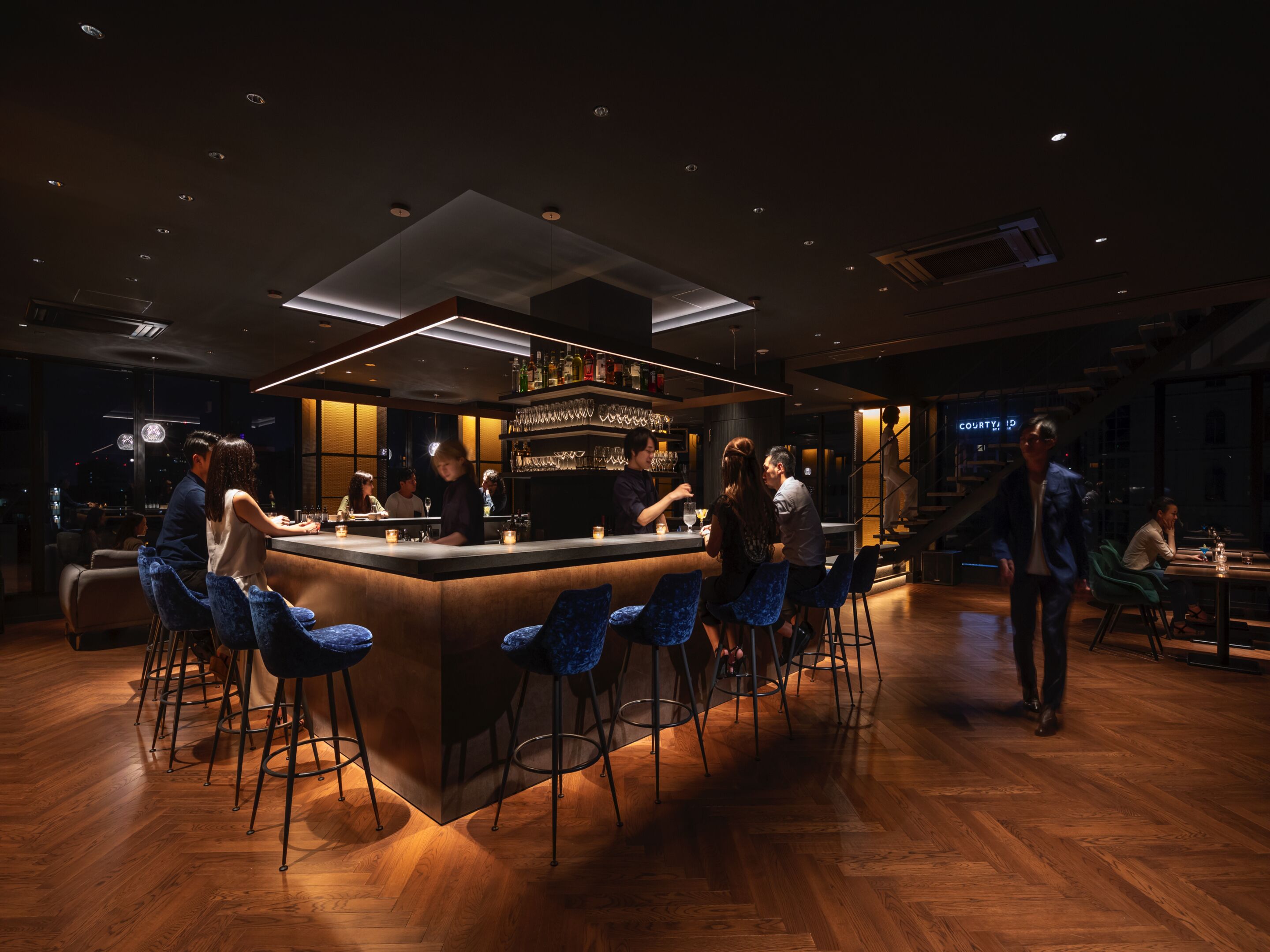 Un bar moderno con una barra central, taburetes y una zona de estar con mesas y sillas.