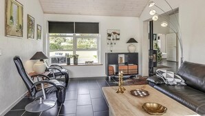A modern living room with a black leather sofa, a wooden coffee table, and a black leather chair. There is a bookshelf with books and a small table with a lamp. A large window provides natural light and a view of greenery outside. - Stunning home in Storvorde with kitchen (Storvorde)