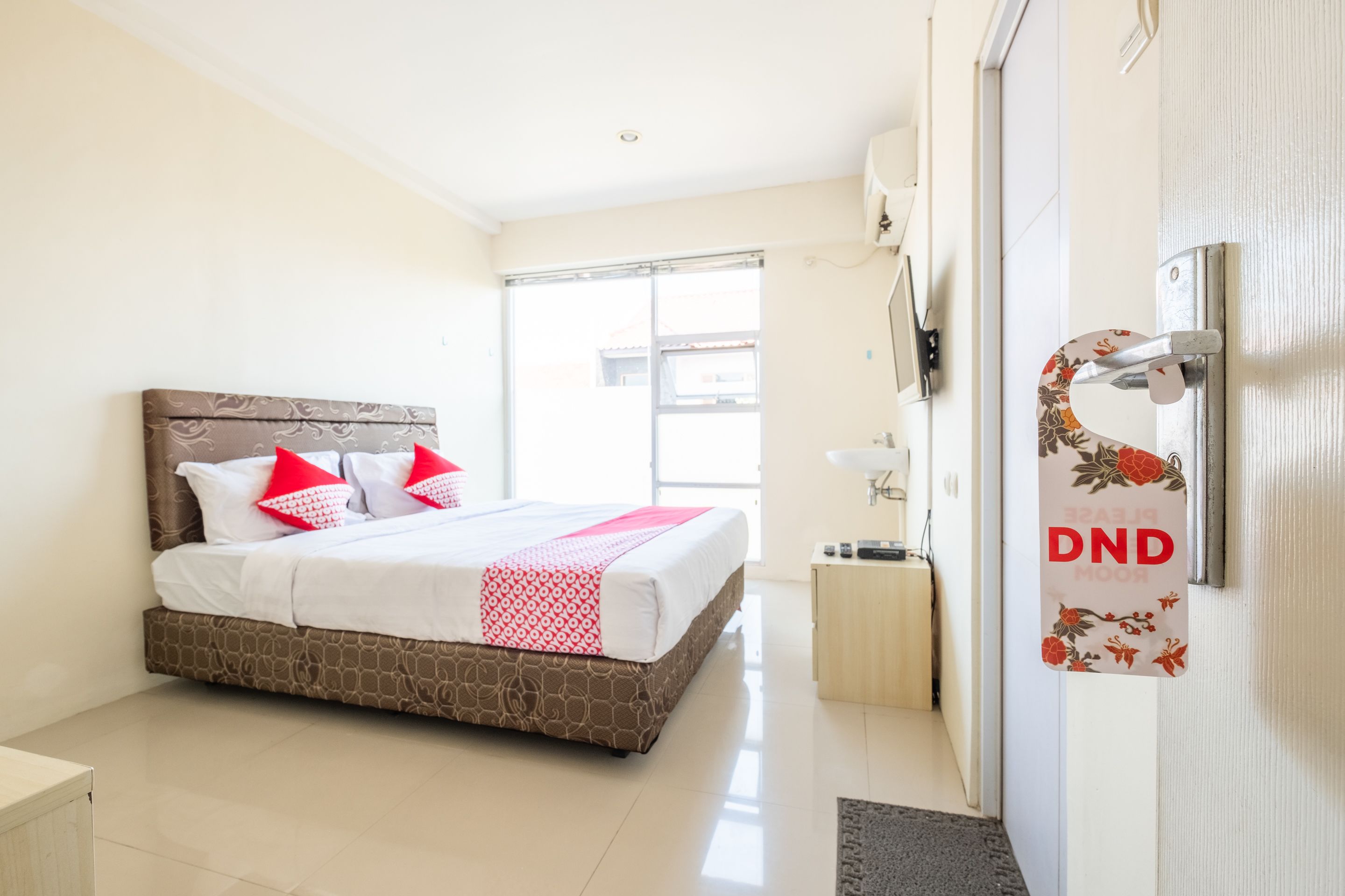 A hotel room with a bed, a bedside table, a nightstand, a window, and a door with a sign.