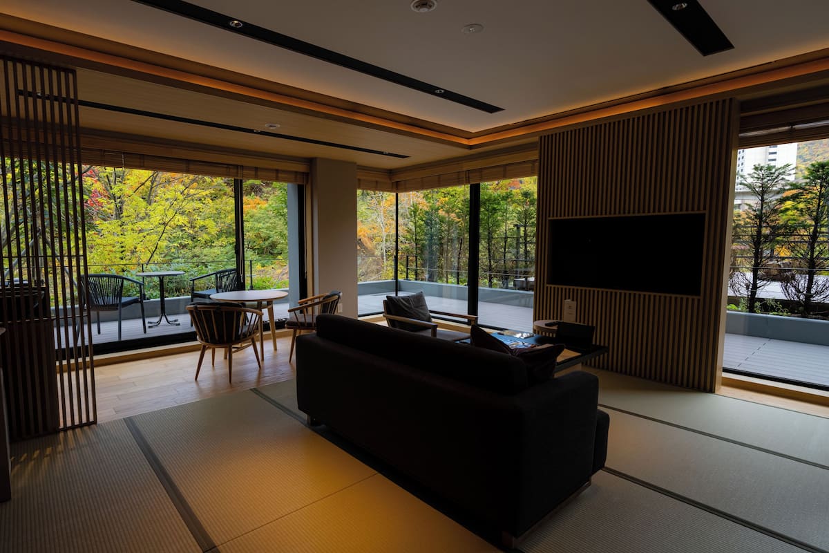 Una sala de estar moderna con sofá, sillas, una mesa de centro y televisor. Amplias ventanas ofrecen vistas a árboles y un estacionamiento.