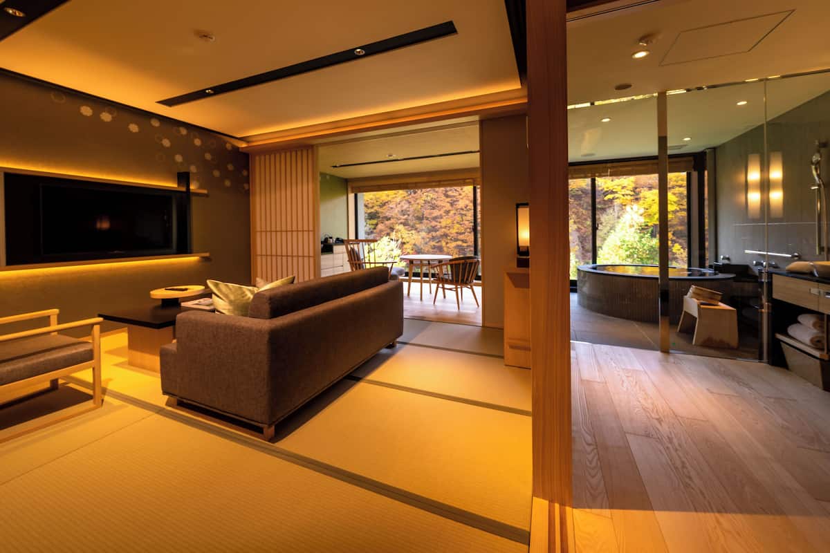 Una sala de estar moderna con un sofá, televisor y un comedor con vistas a árboles otoñales.
