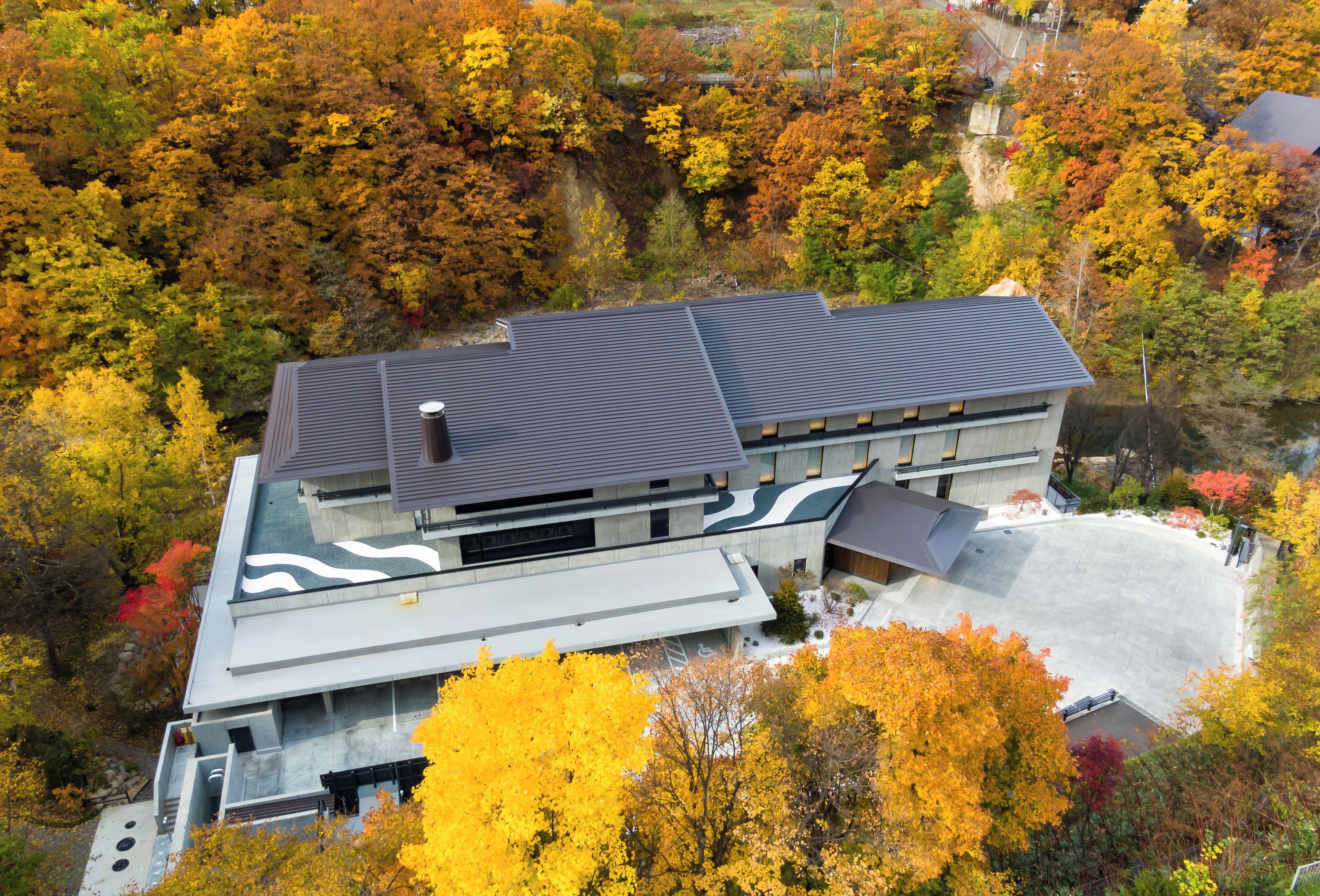 A modern building with a flat roof and a large parking area surrounded by autumn-colored trees.